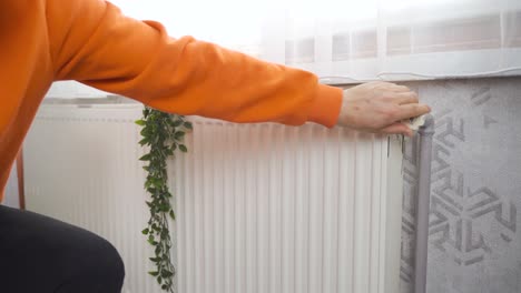 The-man-warms-his-hands-on-the-heating-radiator-by-the-wall.-Cold-in-the-apartment,-poor-heating-system.